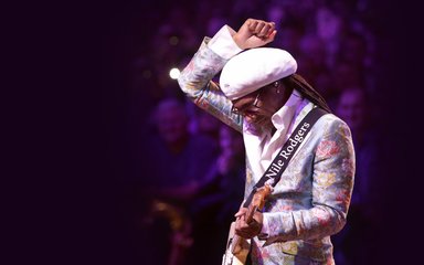 Nile Rodgers performing on stage with one arm in the air and the other arm holding a guitar