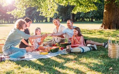 A family of six people sitting around a picnic blanket spread with food, in a woodland setting.
