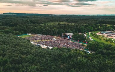 Aerial view of a large crowd in front of a stage, surrounded by forest on all sides.