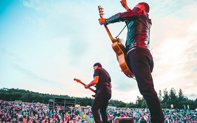 Male singer Bryan Adams, from behind, on stage holding a guitar with a large crowd in view.
