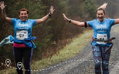 Two smiling women running on a dirt track through woods