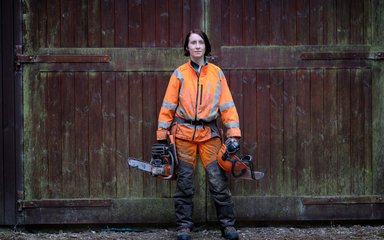 Female forester with chainsaw stood in front of wooden doors 