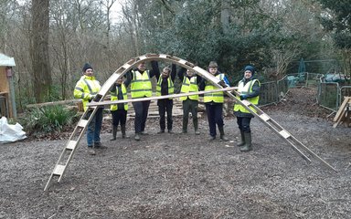 A group of young adults in high vis jackets hold a wooden arch up to pose for a photograph