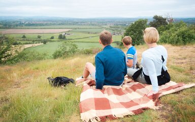 A family having a picnic on a blanket on top of a hill, looking at the view.