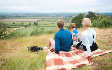 A family having a picnic on a blanket on top of a hill, looking at the view.