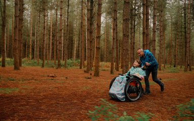 A young girl in a wheelchair smiling as a male carer pushes her through autumn woodland.