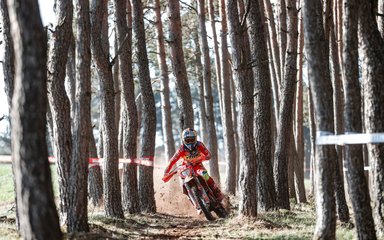 A person in full protective gear rides a motocross bike on a dirt track through a forest