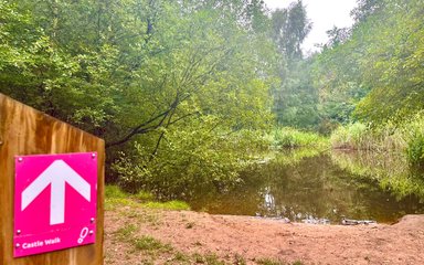 Pink sign with an arrow and the words Castle Walk in front of a pond