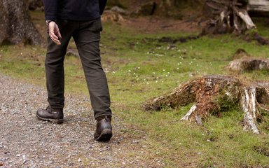 A close-up of a pair of legs walking on a gravel path through a forest.