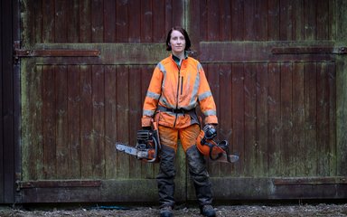 Woman stood in front of large wooden door with helmet and chainsaw in hand 