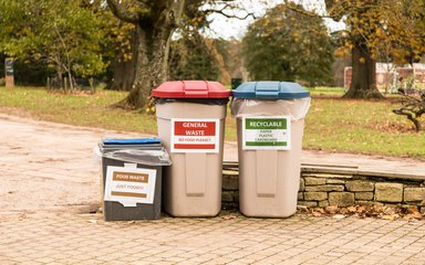 3 Recycling bins stand side by side. A shorter brown box asks for food waste, a taller light brown bin with a red lid says general waste and a same height light brown bin with a blue lid says Recyclable. 