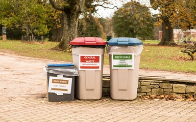 3 Recycling bins stand side by side. A shorter brown box asks for food waste, a taller light brown bin with a red lid says general waste and a same height light brown bin with a blue lid says Recyclable.