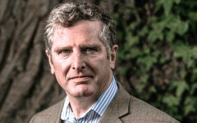 Head-and-shoulders portrait of John Varley, wearing a brown blazer and striped shirt outdoors.