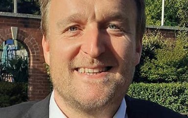 A man with light blonde hair and a light stubbly beard smiles at the camera