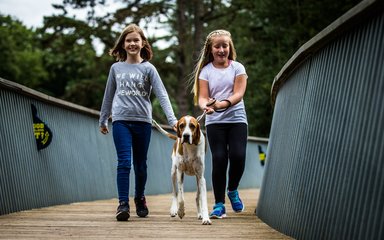 2 young girls with a dog on a lead walk over a wooden walkway with steel railings 