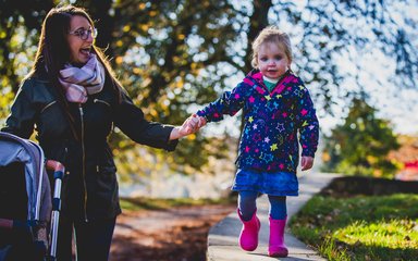 A toddler in a bright blue starry coat and pink welly boots walks along holding her mums hand over a flat low wall. The mum in a green coat and pink tartan scarf cheers and encourages her girl on