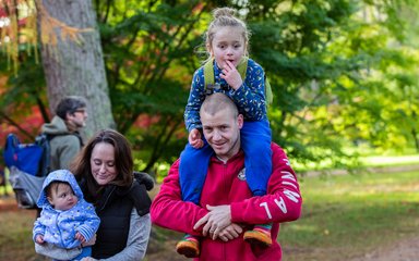 A young boy sits on his fathers shoulders. His mum walks beside them holding a baby.