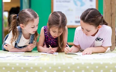 three young girls lean on a table to write and draw on paper together.
