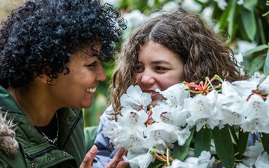 A mum and daughter smile at each other while smelling white rhododendron flowers