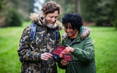 An adult male and female dressed in green coats look at a Friends of Westonbirt Arboretum leaflet while smiling at the great membership scheme.