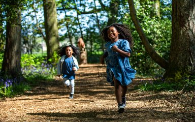 2 children run towards the camera smiling - they are running between trees with a sculpture of an owl in the background