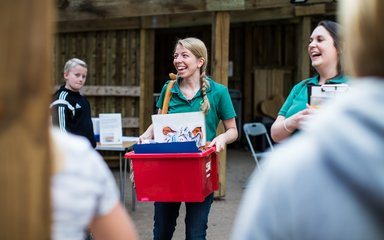An adult female holds a bright red box full of learning activities for the day. Beside her is another adult female holding a clipboard. They both laugh as they welcome a group of children to the arboretum