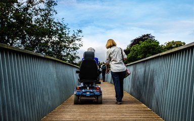 Two women; one walking and one on an electric scooter on a bridge with trees and blue sky