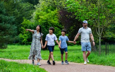two boys, a man and a woman holding hands in a line on a path with grass and trees either side. The woman is pointing.