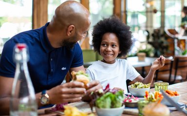 A father and son sit at a table with food in front of them. The father looks at his son who smiles back.