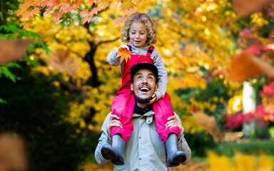A boy sits on his dad shoulders holding a yellow maple leaf surrounded by autumn colour