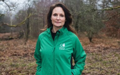 A woman with long dark hair, wearing a green jacket that says 'Forestry England', standing in woodland.