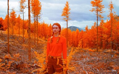 A red-haired woman wearing orange standing near trees with orange foliage.