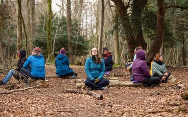 A group of people in coats sit in a circle on the forest floor