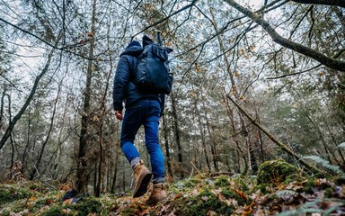 A man with his back to the camera, wearing walking boots, jeans, coat and hat, walking through the forest in winter.