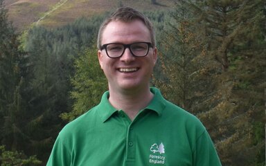 A white man with short hair and black-rimmed glasses, wearing a green Forestry England t-shirt.
