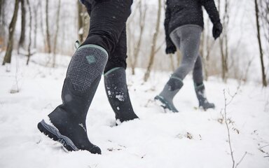 Two pairs of legs wearing hardy rubber boots in the snow.