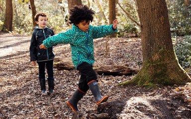 Two children in forest, one in coat and wellies is running over a dirt mound