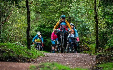 A group of riders cycling in a forest, led by a woman with a baby