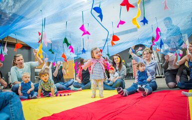 Image of a group of parents and small children playing under blue fabric with mobiles hanging