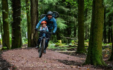 A smiling man and child on a mountain bike in the forest.