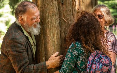 An older man and two older women smiling at each other and hugging a large tree trunk