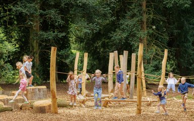 A wooden playground featuring ropes and wooden stumps to climb on. Children are having fun playing.
