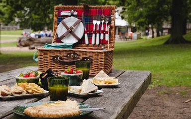 A wicker hamper with red tartan lining on a forest picnic table, shown with plates of good and drinks glasses.