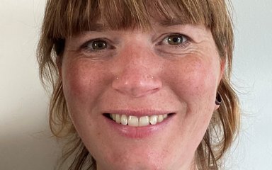 Portrait photo of a woman with light brown hair tied back with a fringe, brown eyes and a friendly smile.