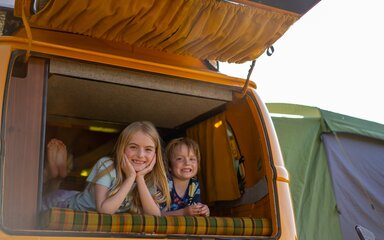 A boy and a girl smiling as they look out the back of an orange campervan