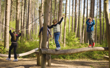 4 children on a wooden balance beam with a rope for them to hold on to