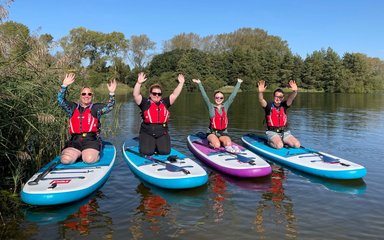 Four people kneeling on paddle boards afloat on water, smiling with their hands in the air.
