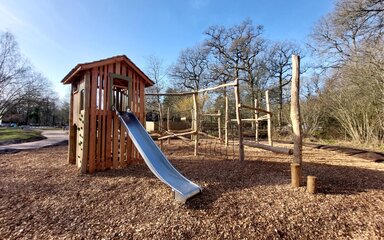 Metal slide coming from wooden play tower with climbing nets and balance beams