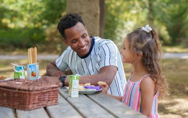 A father and daughter smiling at each other whilst sat at an outdoor picnic bench.