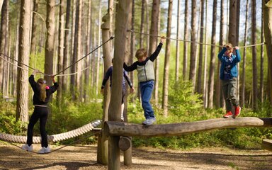 4 children on a wooden balance beam with a rope for them to hold onto.